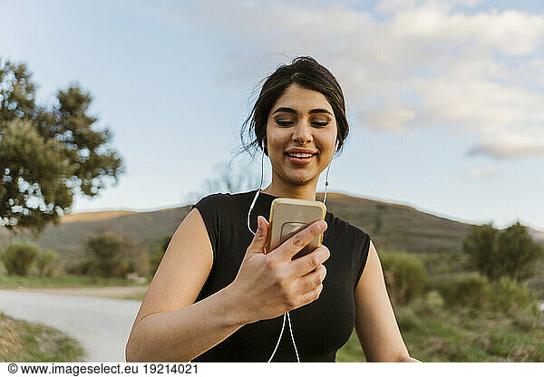 Smiling woman holding mobile phone and exercising on footpath