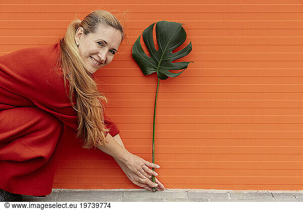 Smiling woman holding monstera leaf and crouching in front of orange wall
