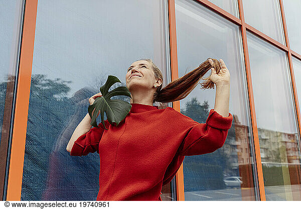 Smiling woman holding monstera leaf and pulling ponytail in front of building
