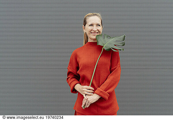 Smiling woman holding monstera leaf and standing in front of gray wall