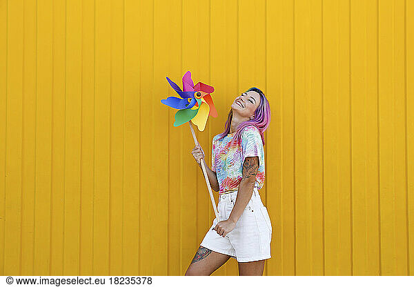Smiling woman holding multi colored pinwheel toy in front of yellow wall
