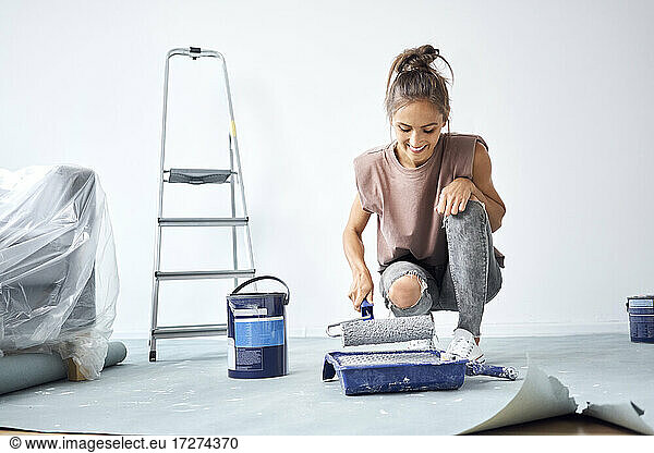 Smiling woman holding paint roller while crouching at home