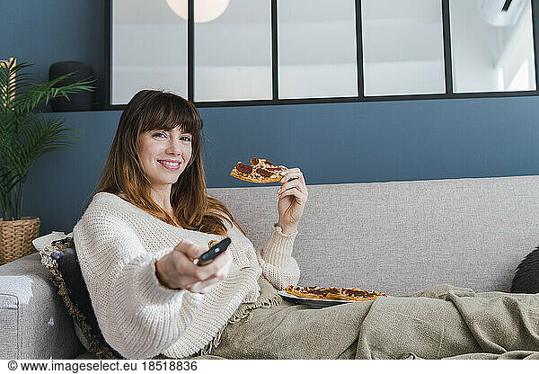Smiling woman holding pizza and watching TV sitting on sofa at home