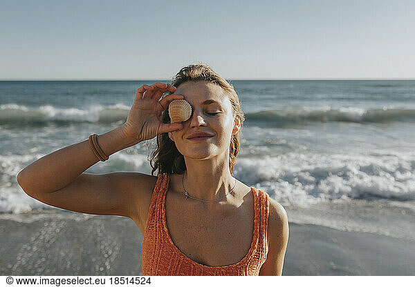 Smiling woman holding seashell over eye in front of sea