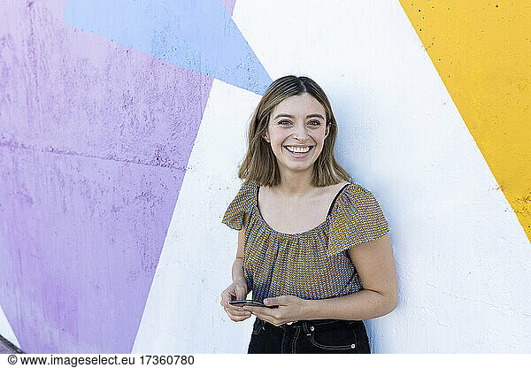Smiling woman holding smart phone in front of colorful wall