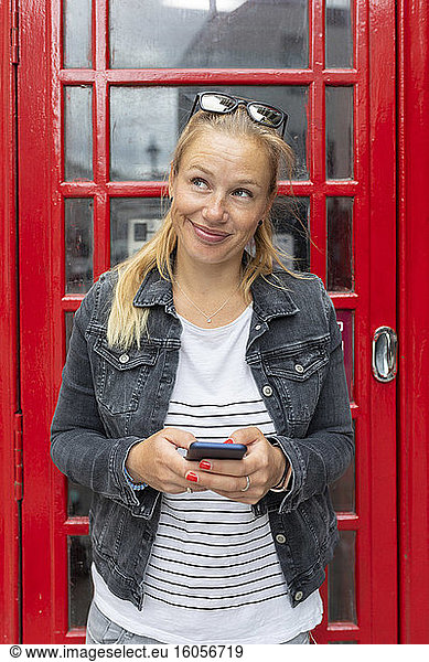 Smiling woman holding smart phone looking away while standing against telephone booth