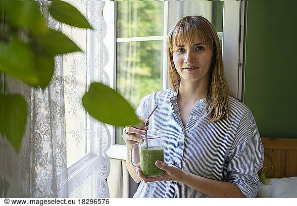 Smiling woman holding smoothie standing by window
