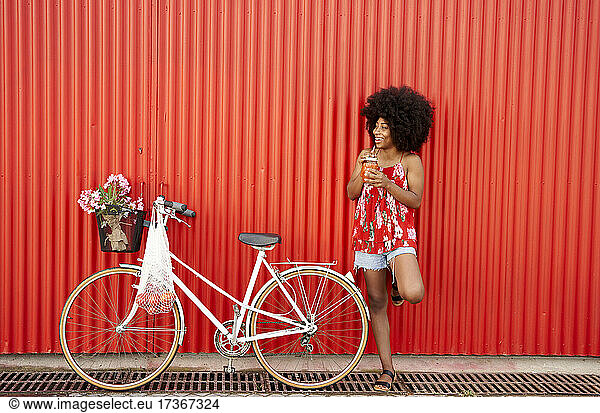 Smiling woman holding smoothie while standing by bicycle in front of red wall