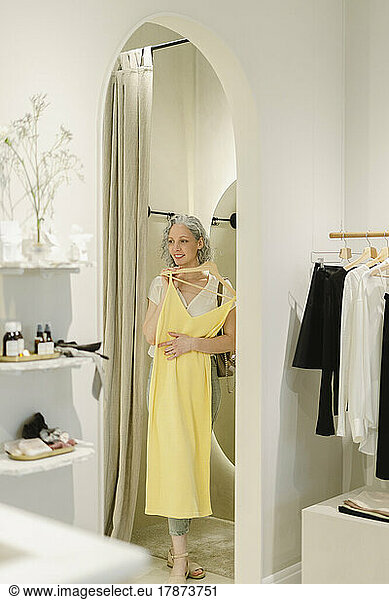 Smiling woman holding yellow dress stepping out from dressing room at store