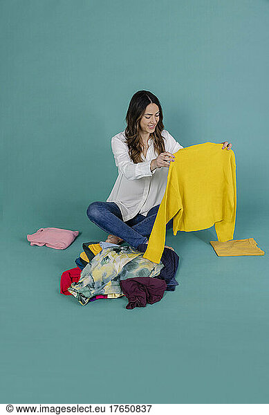 Smiling woman holding yellow jumper sitting with clothes against blue background