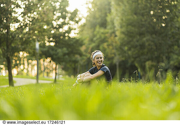 Smiling woman hugging knees while sitting on grass at public park