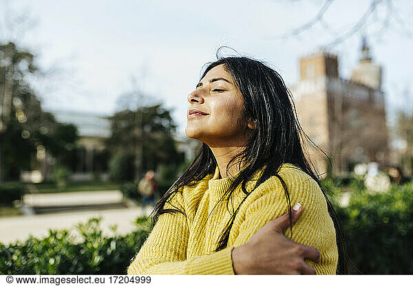 Smiling woman hugging self against sky in public park