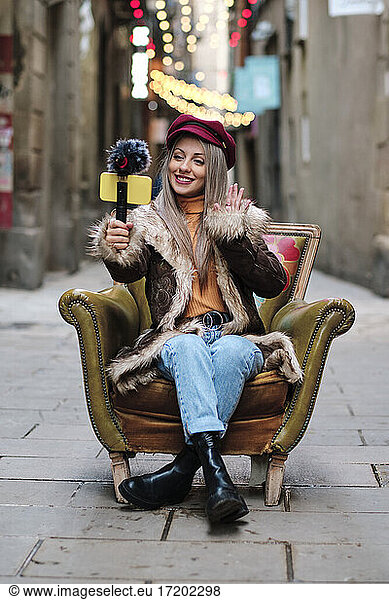Smiling woman in fur coat vlogging through smart phone while sitting on armchair at alley