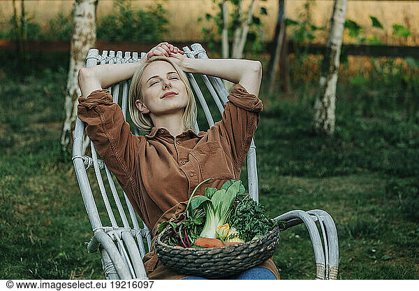 Smiling woman leaning on chair with vegetable basket in garden