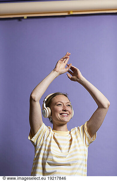 Smiling woman listening music on headphones and dancing over purple background