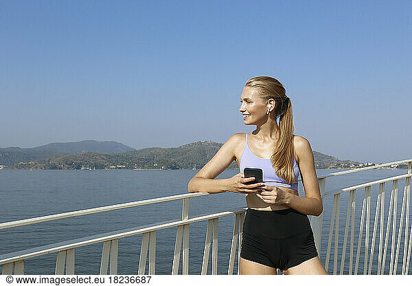 Smiling woman listening to music through wireless in-ear headphones holding mobile phone leaning on railing