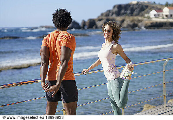 Smiling woman looking at boyfriend doing stretching exercise on pier in coastal area