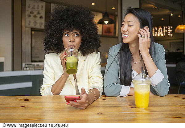 Smiling woman looking at friend drinking juice in cafe