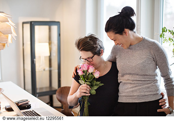 Smiling woman looking at girlfriend smelling pink roses at home