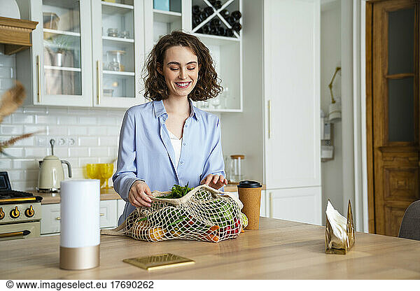 Smiling woman looking at vegetables and fruits in mesh bag at home