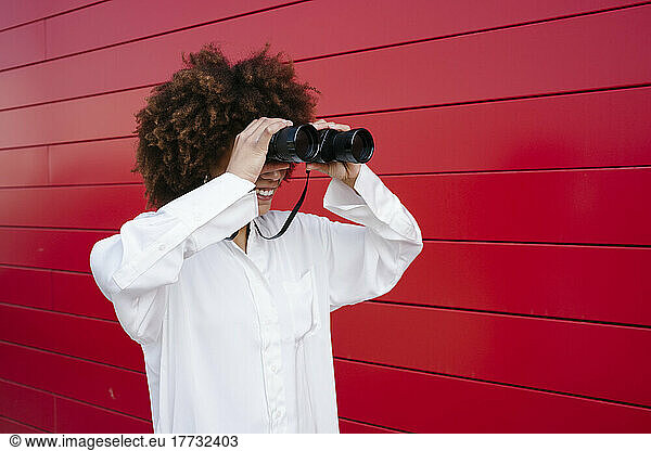 Smiling woman looking through binoculars by red wall