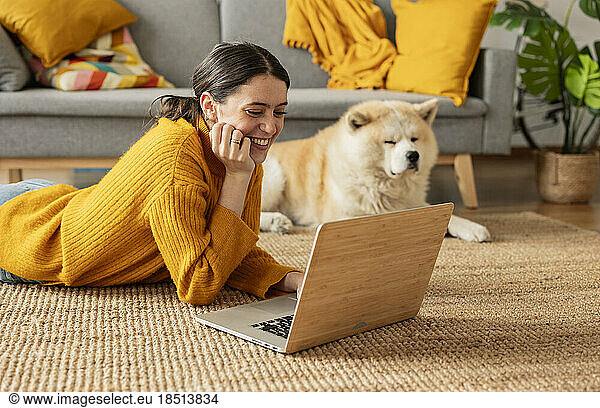 Smiling woman lying on carpet and using laptop by dog at home