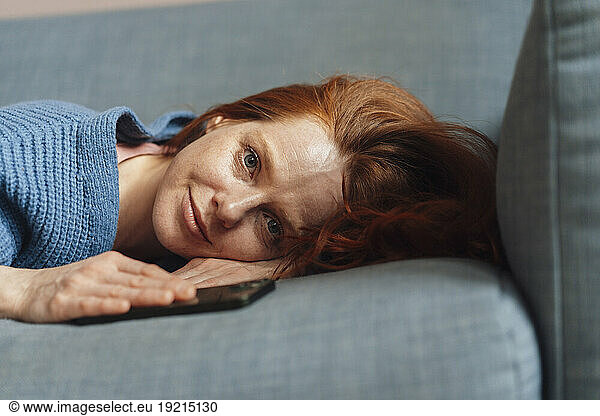 Smiling woman lying on gray sofa with smart phone at home