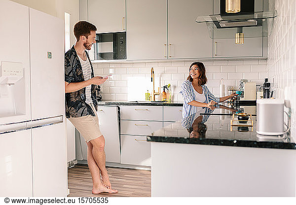 Smiling woman making coffee while looking at man using phone in kitchen