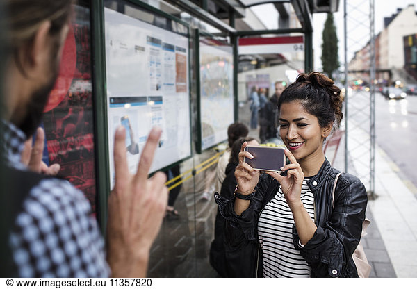 Smiling woman photographing man through smart phone at bus stop in city