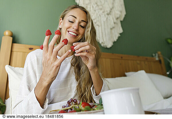 Smiling woman putting raspberries on fingers at home