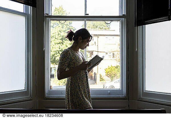 Smiling woman reading book standing in front of window