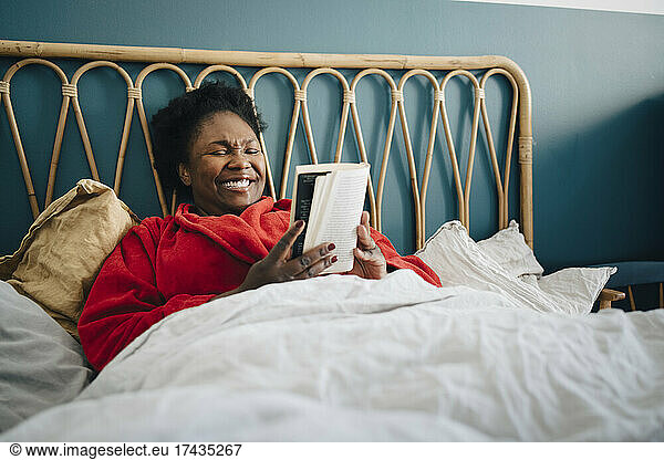 Smiling woman reading book while sitting on bed at home