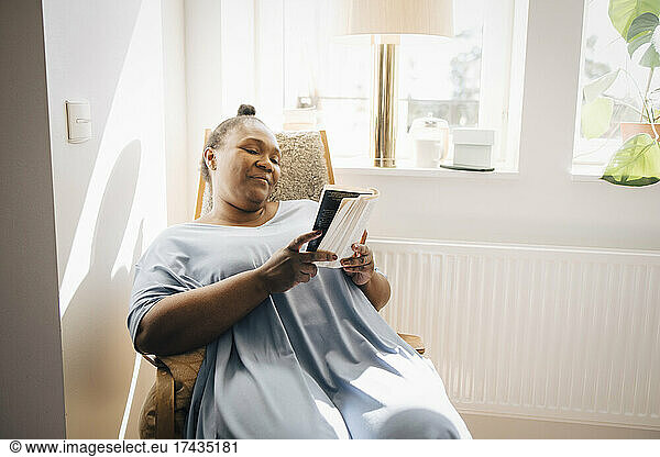 Smiling woman reading book while sitting on chair in living room