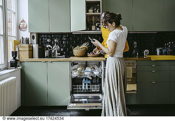 Smiling woman sharing smart phone to boyfriend doing dishes in kitchen at home