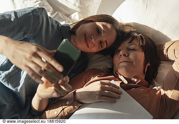 Smiling woman sharing smart phone with lesbian friend lying down at home