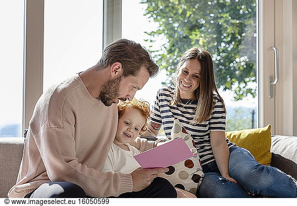 Smiling woman sitting and looking at man reading picture book to boy in living room