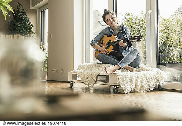 Smiling woman sitting at the window at home playing guitar