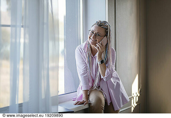Smiling woman sitting by window and talking on smart phone at home