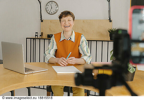 Smiling woman sitting in classroom looking at camera recording online class