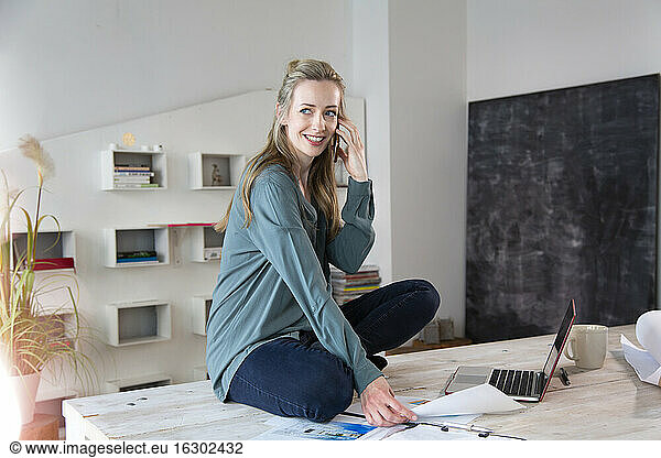 Smiling woman sitting on desk in home office talking on the phone
