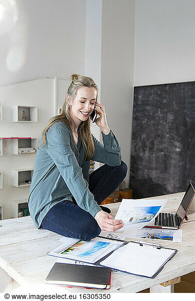 Smiling woman sitting on desk in home office talking on the phone