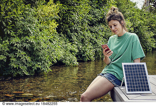 Smiling woman sitting on patio using smart phone getting charged by solar panel