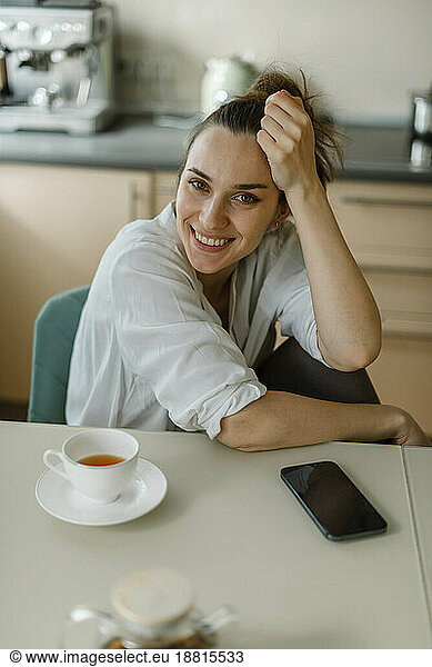 Smiling woman sitting on table with smart phone and tea
