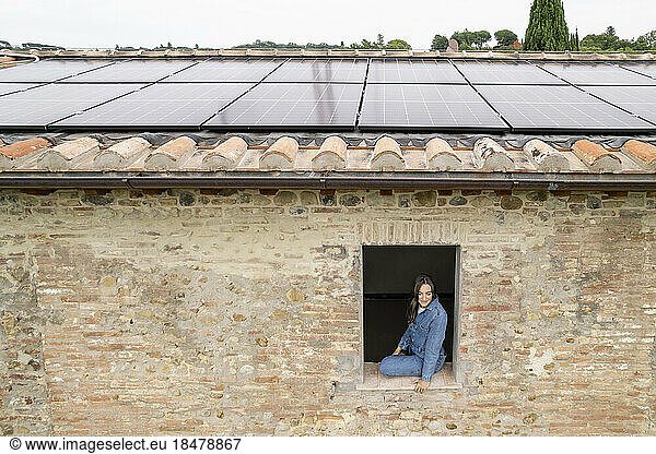 Smiling woman sitting on window sill with solar panels on rooftop