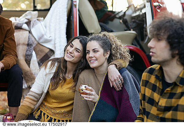 Smiling woman sitting with arm around friend on picnic outside campervan