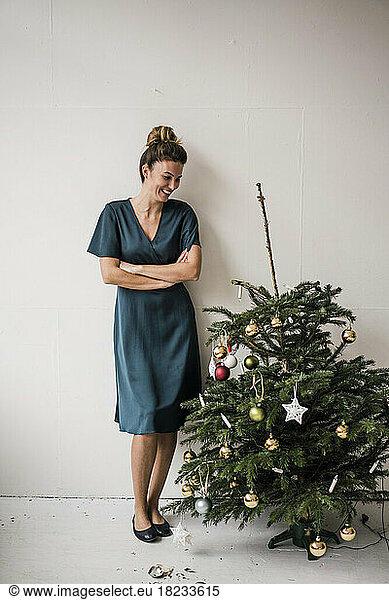 Smiling woman standing with arms crossed by damaged Christmas tree in front of white wall