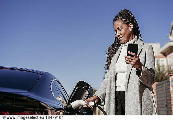 Smiling woman standing with smart phone and charging car on sunny day