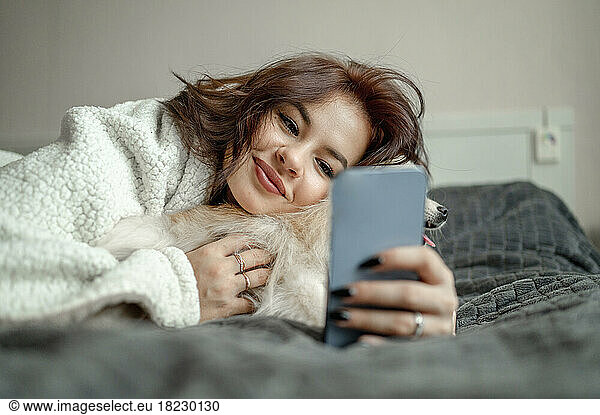 Smiling woman stroking Pomeranian dog and using smart phone at home