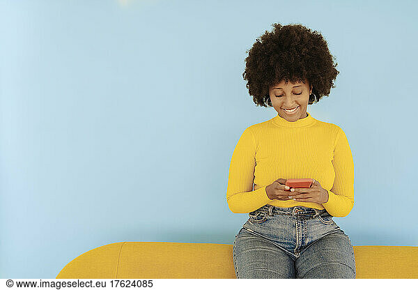 Smiling woman surfing net through mobile phone sitting on yellow sofa against blue background