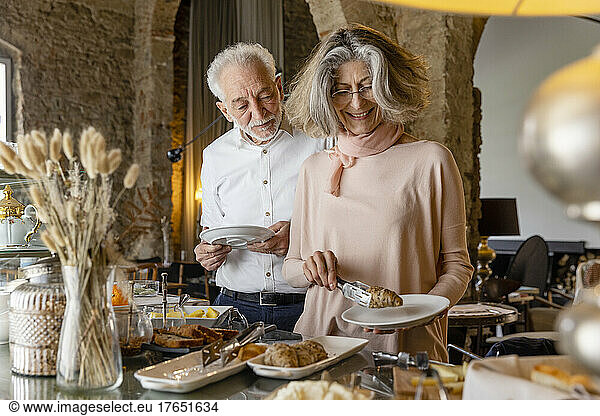 Smiling woman taking croissant from buffet standing by man at boutique hotel
